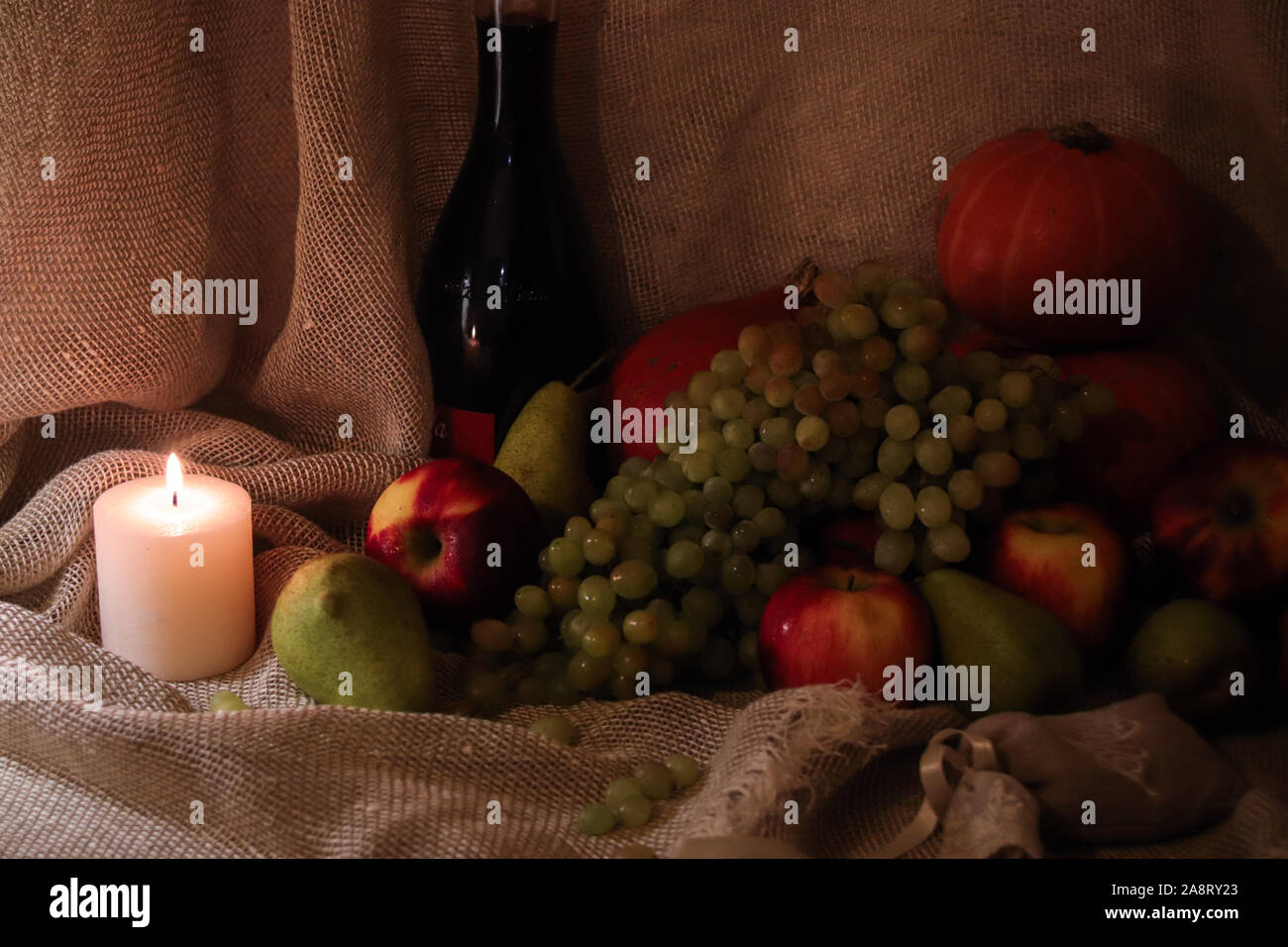 Autumn harvest still life. Apples, pears, grapes and pumpkin on sackcloth backgound with bottle of wine and candle light Stock Photo