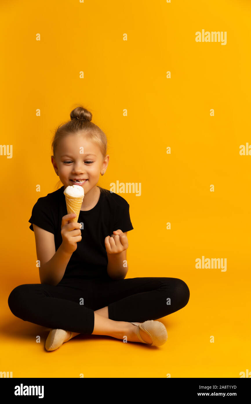 Beautiful little girl child bites vanilla ice cream in a waffle cone on a yellow background Stock Photo