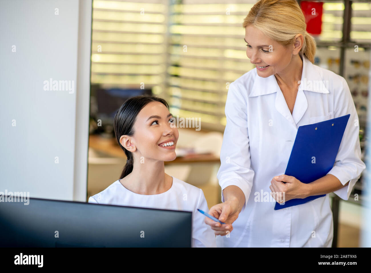 Blonde eye doctor smiling while talking to her pleasant trainee Stock Photo