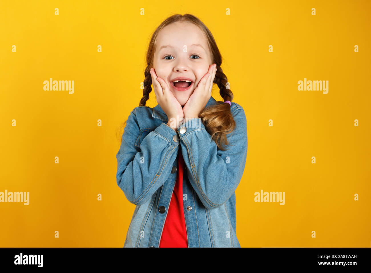 Cheerful emotional little girl in a denim shirt on a yellow background. The child is pleased with the good news. Stock Photo