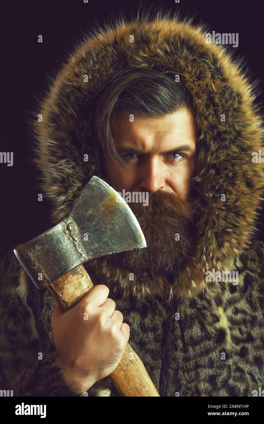 handsome bearded man or woodman guy with fashionable mustache and beard on serious face in brown fur coat, holds sharp axe or ax on black background Stock Photo