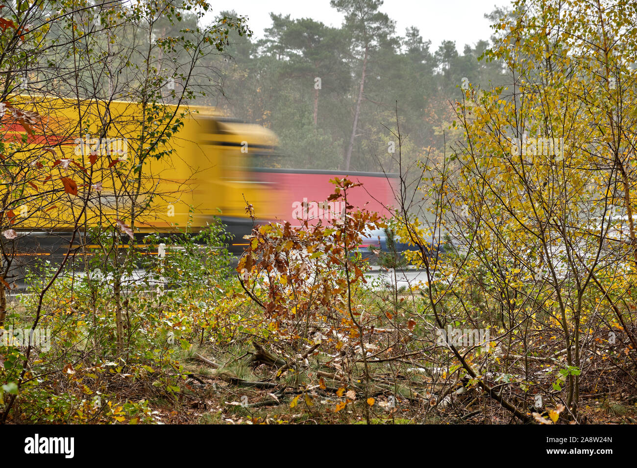 Trucks driving with speed along the highway behind some small autumn trees and bushes. Seen in Bavaria, Germany on a wet and rainy autumn day. Stock Photo