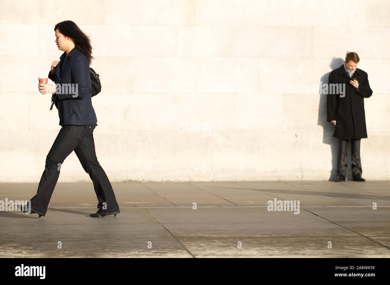 LONDON - NOVEMBER 17, 2011: A woman carrying a coffee cup passes a man standing looking at his mobile phone in Trafalgar Square. Stock Photo