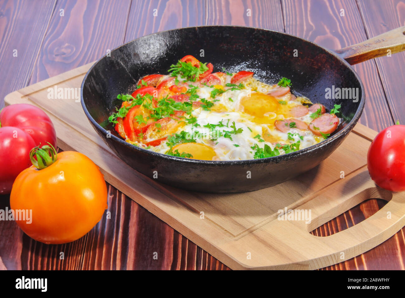 Scrambled eggs with tomatoes, sausages, sweet pepper and sprinkled with chopped parsley in a frying pan Stock Photo