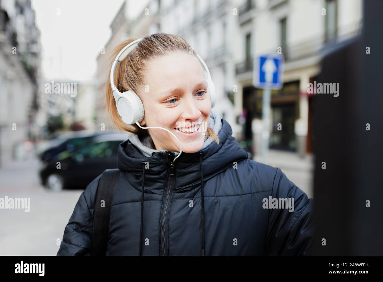 Young blonde woman paying on parking meter in the city with surprised face wearing a jacket an headphones Stock Photo