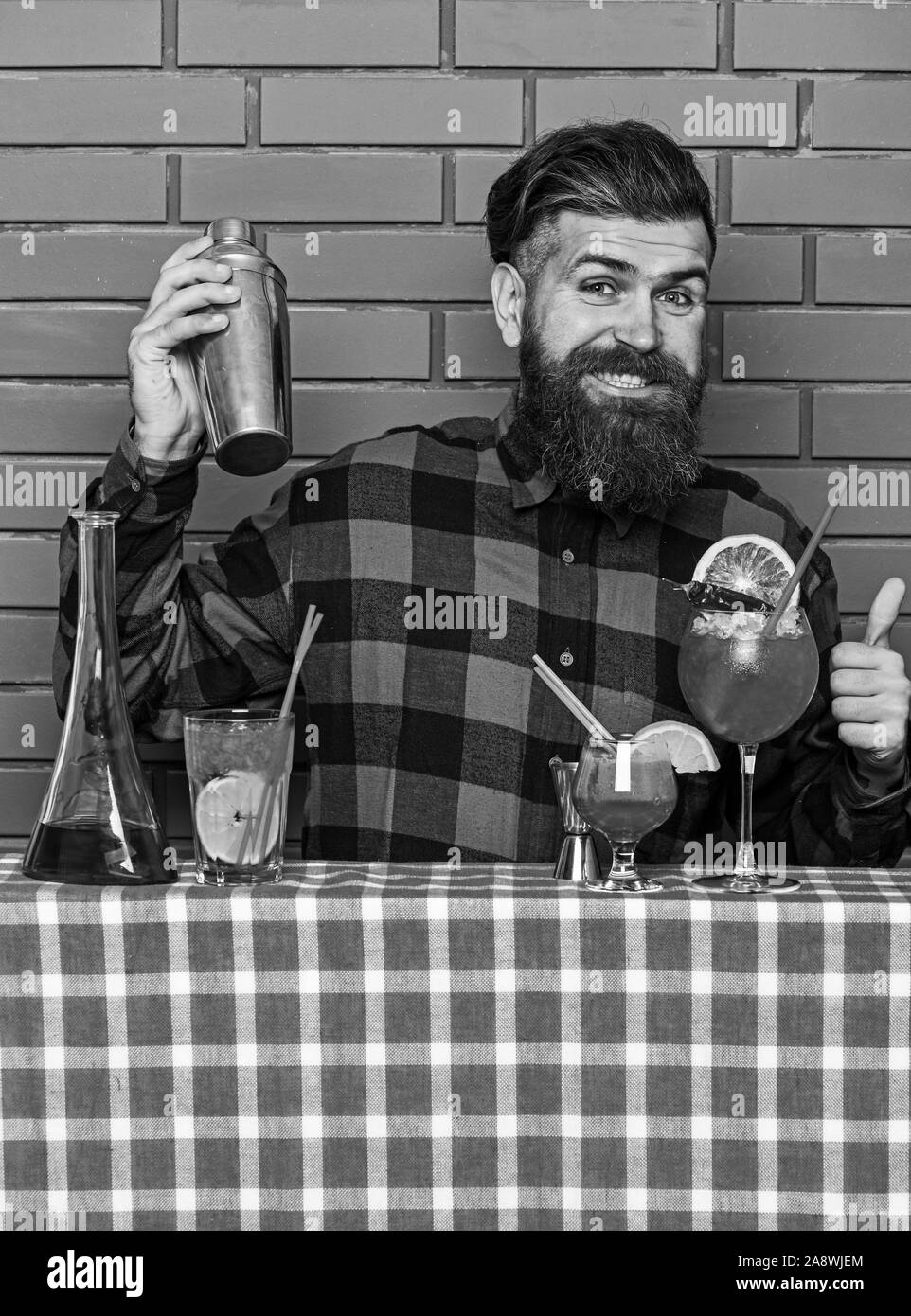 Alcoholic drinks concept. Man in checkered shirt on brick wall background prepares drinks. Barman with long beard and mustache and stylish hair on happy face holding shaker, makes alcoholic cocktail. Stock Photo