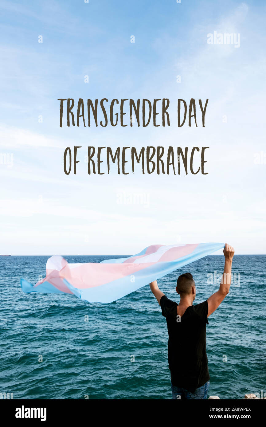 a young caucasian person, seen from behind, in front of the sea holding a transgender pride flag on the air and the text transgender day of remembranc Stock Photo