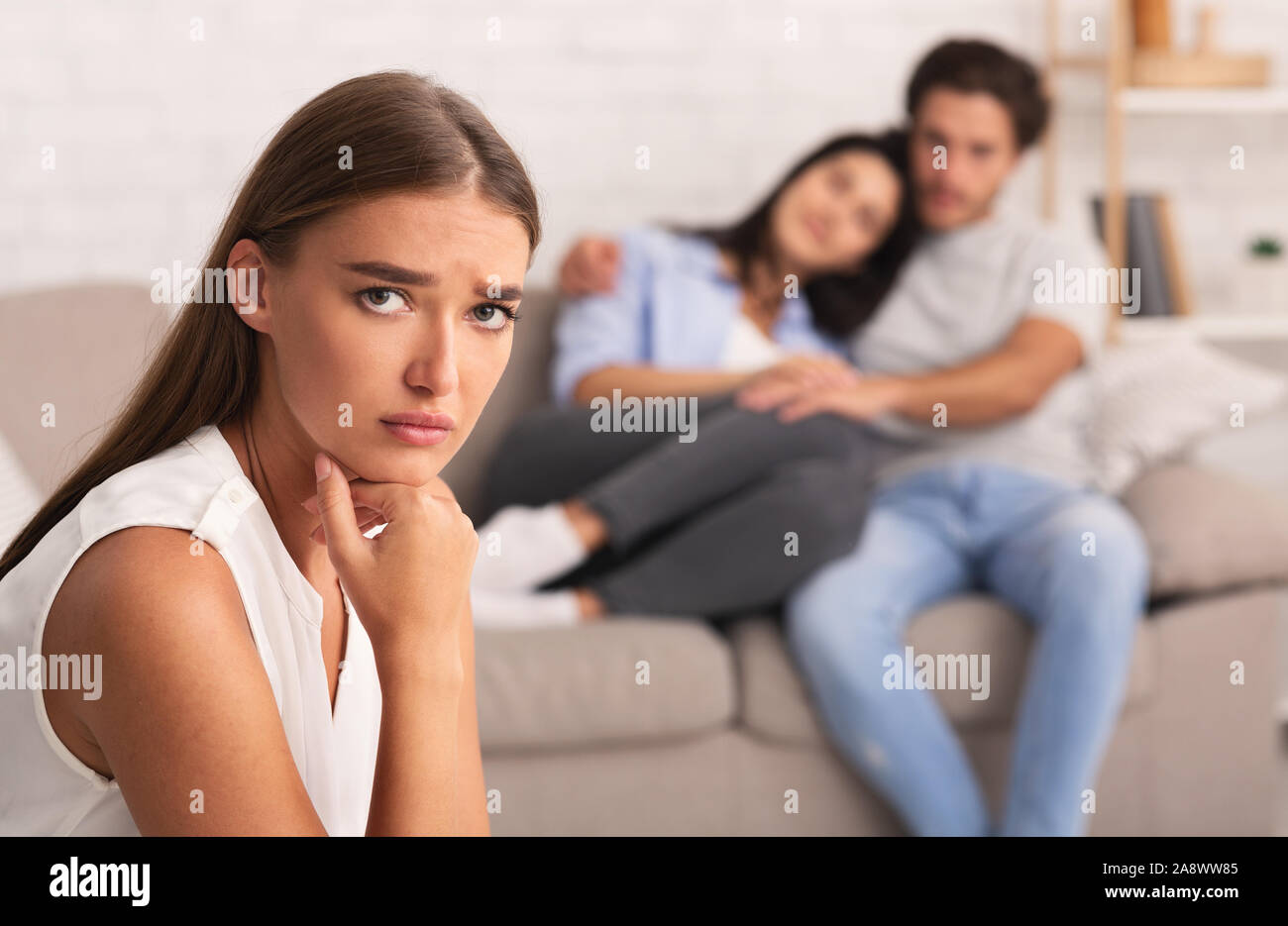 Unhappy Girl Looking At Camera While Her Friends Hugging Indoor Stock Photo