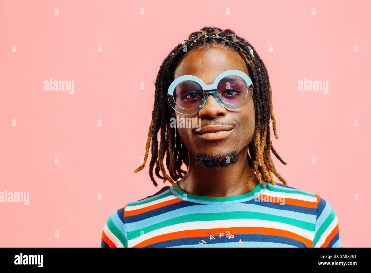 Portrait of a smiling young man in striped shirt with 'happy' sign and big sunglasses, standing in front of a pink background Stock Photo