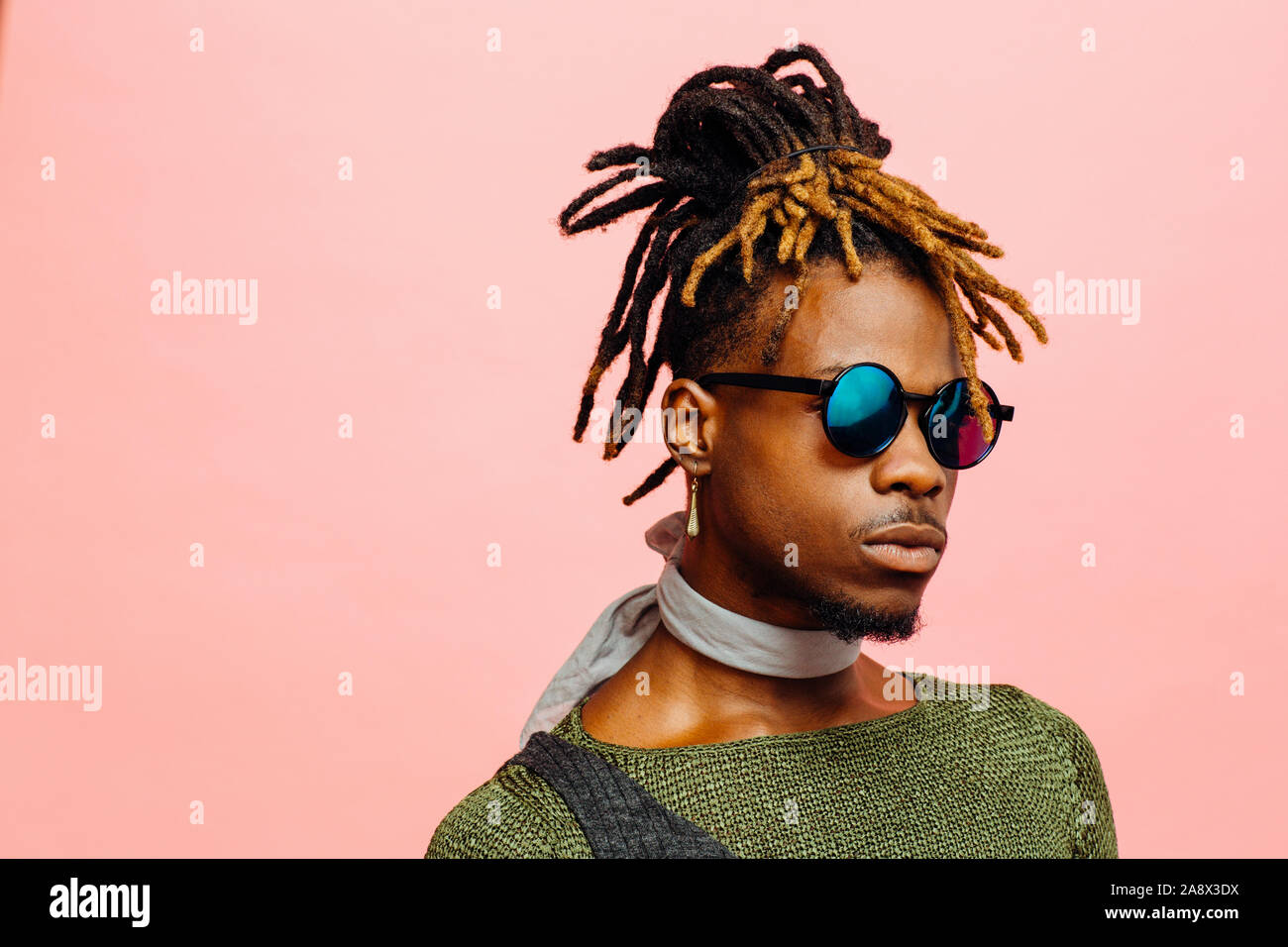 Close up portrait of a young man in green with dreadlocks and blue sunglasses, isolated on pink Stock Photo