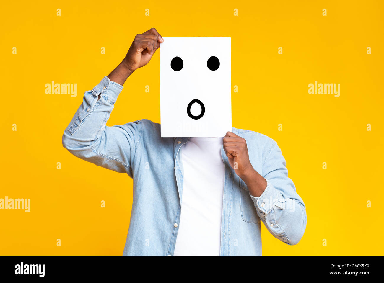 Black guy hiding face behind paper with drawn surprised emoticon Stock Photo
