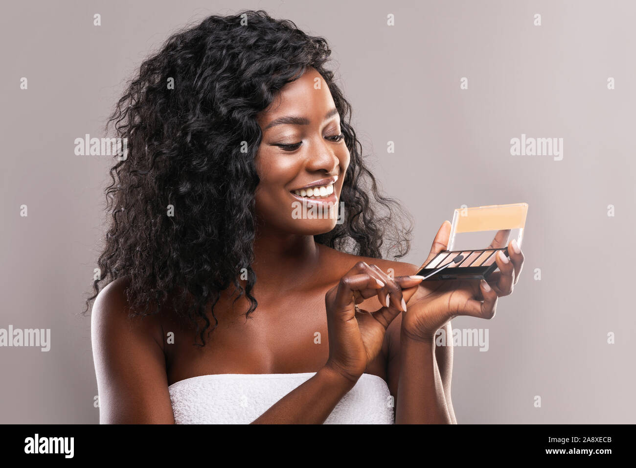 Beautiful black girl trying new eyeshadow palette over gray background Stock Photo