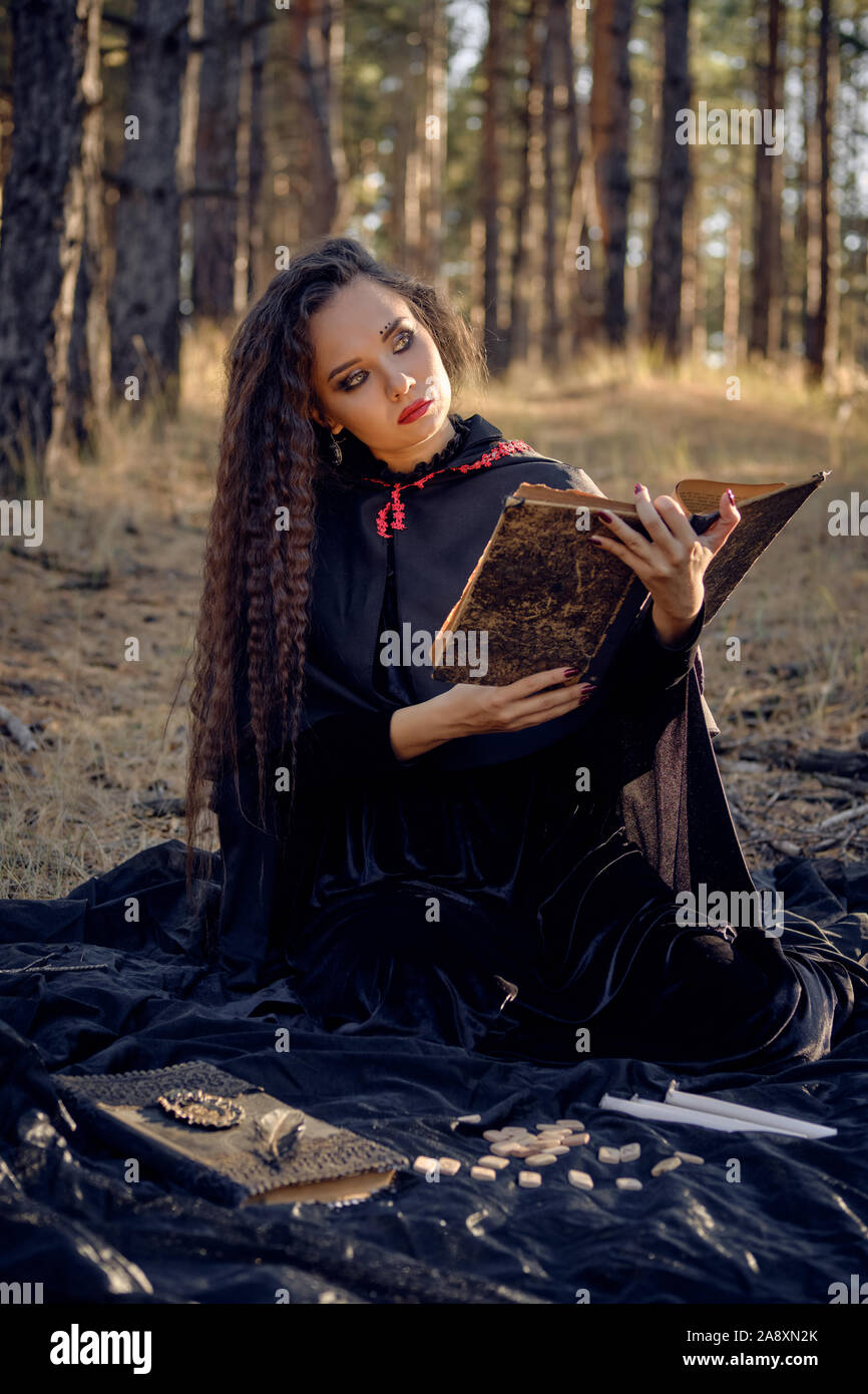 Pretty, wicked, long-haired sorceress in a black, long dress with cape and hood. She is reading a book, sitting on a dark blanket whith runes and cand Stock Photo