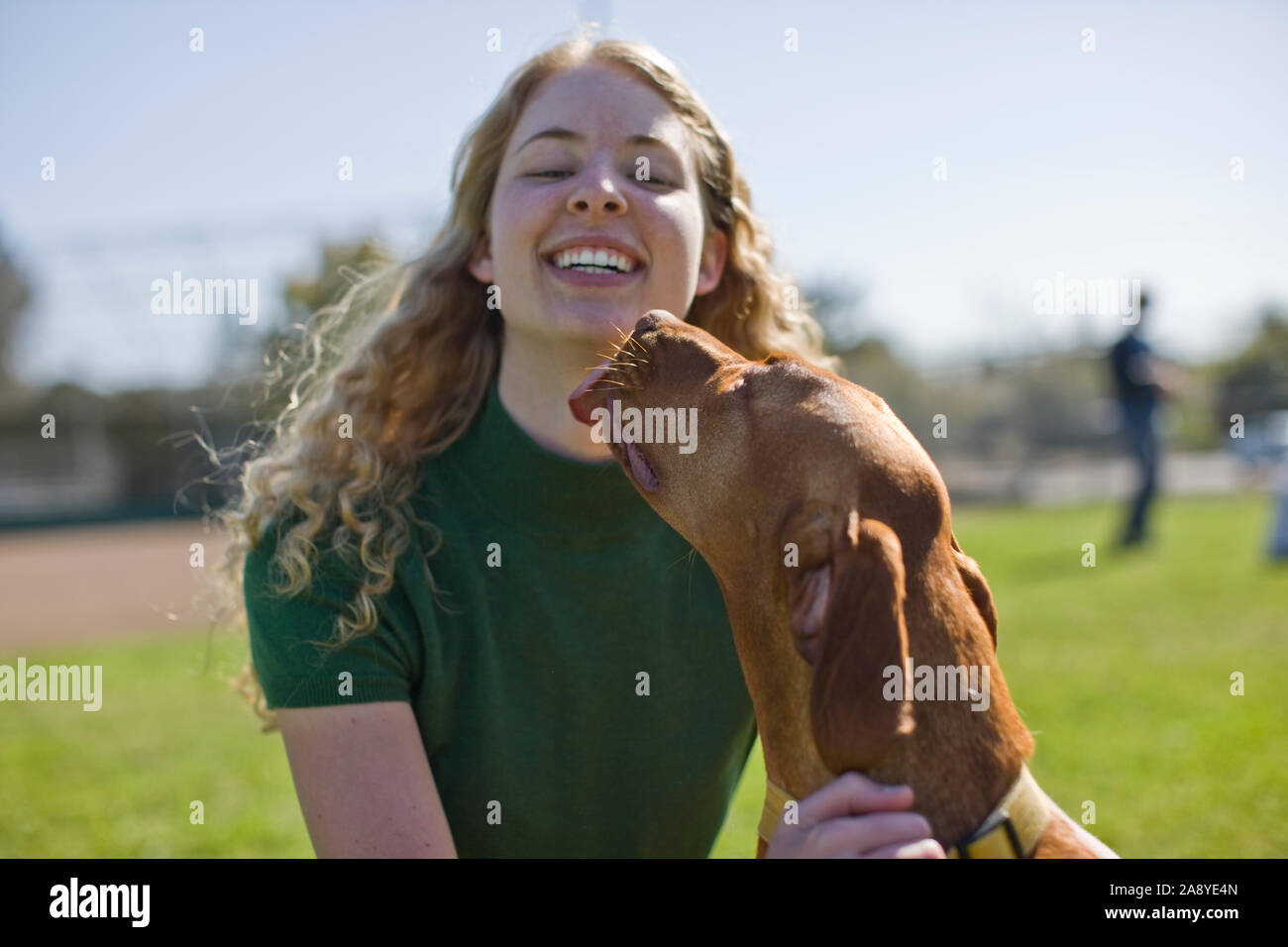 Smiling teenage girl being licked on the chin by her brown dog while at the park. Stock Photo