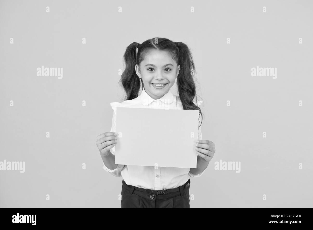 Here is your homework. Adorable small child holding empty homework sheet on yellow background. Cute little girl with blank green school paper for doing homework assignment. Homework, copy space. Stock Photo