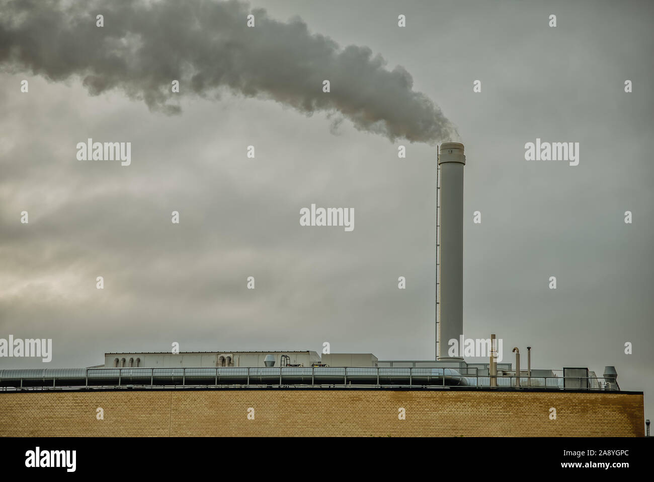 Black smoke on a grey sky, comming from a tall chimney on the top of a factory. Denmark, November 10, 2019 Stock Photo