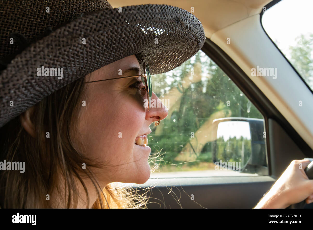 Selective focus on head of happy young woman driving her SUV car while wearing a big hat and funky sunglasses. Having good time while traveling. Stock Photo