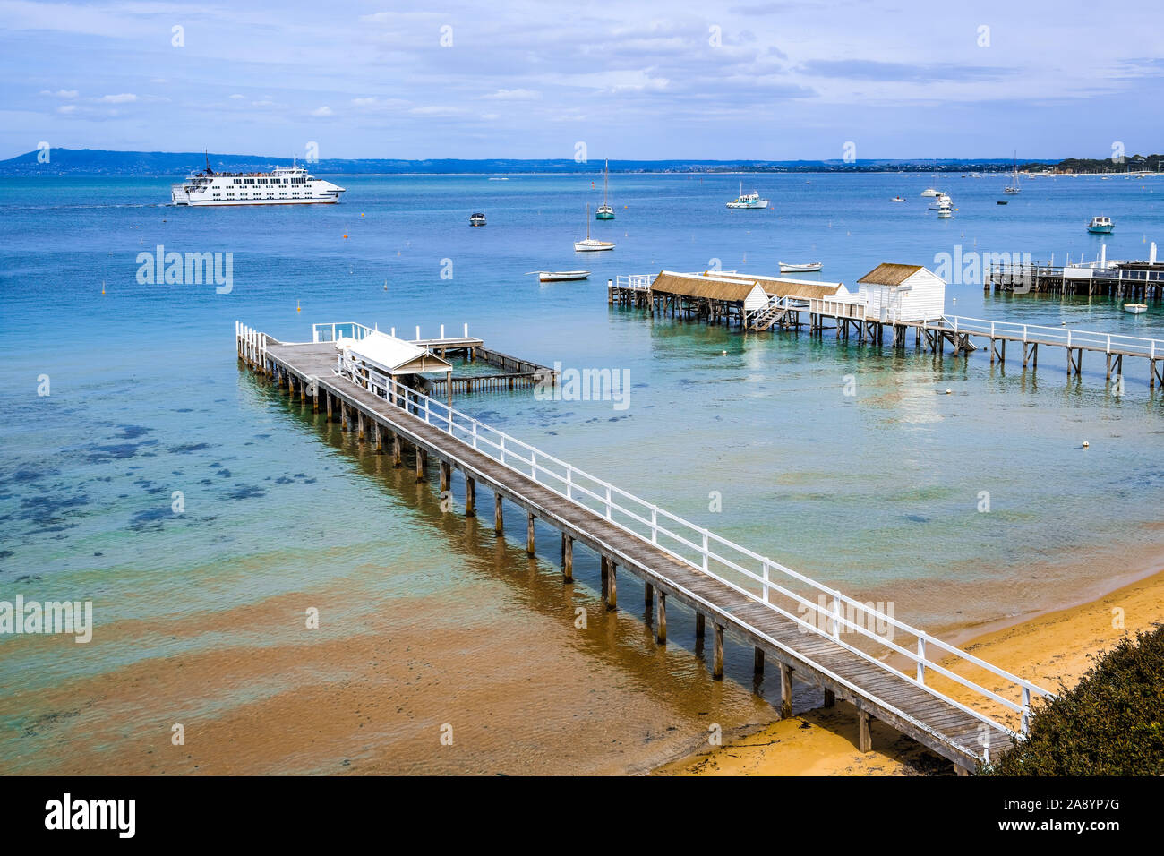 Coastal landscape - wooden piers extending into shallow bay water with passenger ferry sailing in the distance. Melbourne, Victoria, Australia Stock Photo
