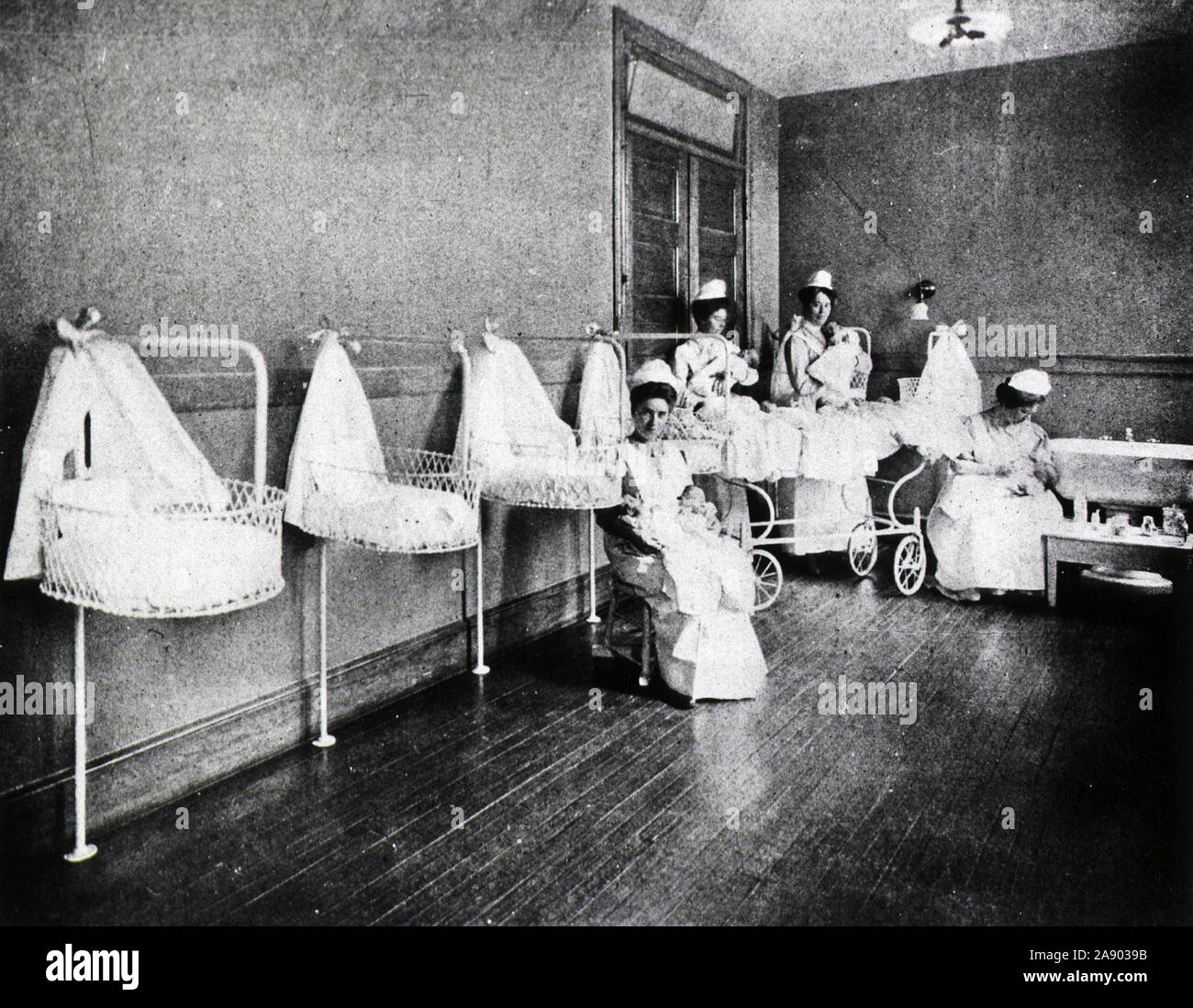 Four nurses hold infants in a large room with baby baskets mounted along one wall. Stock Photo