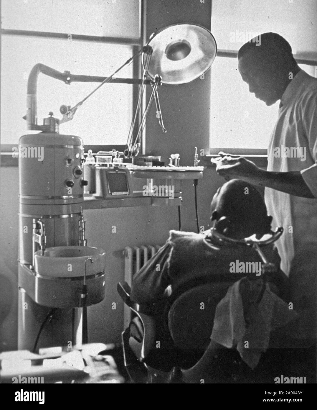 Interior view of a dental office with an African American patient sitting in the dental chair and a dentist standing next to him ca. 1947 Stock Photo