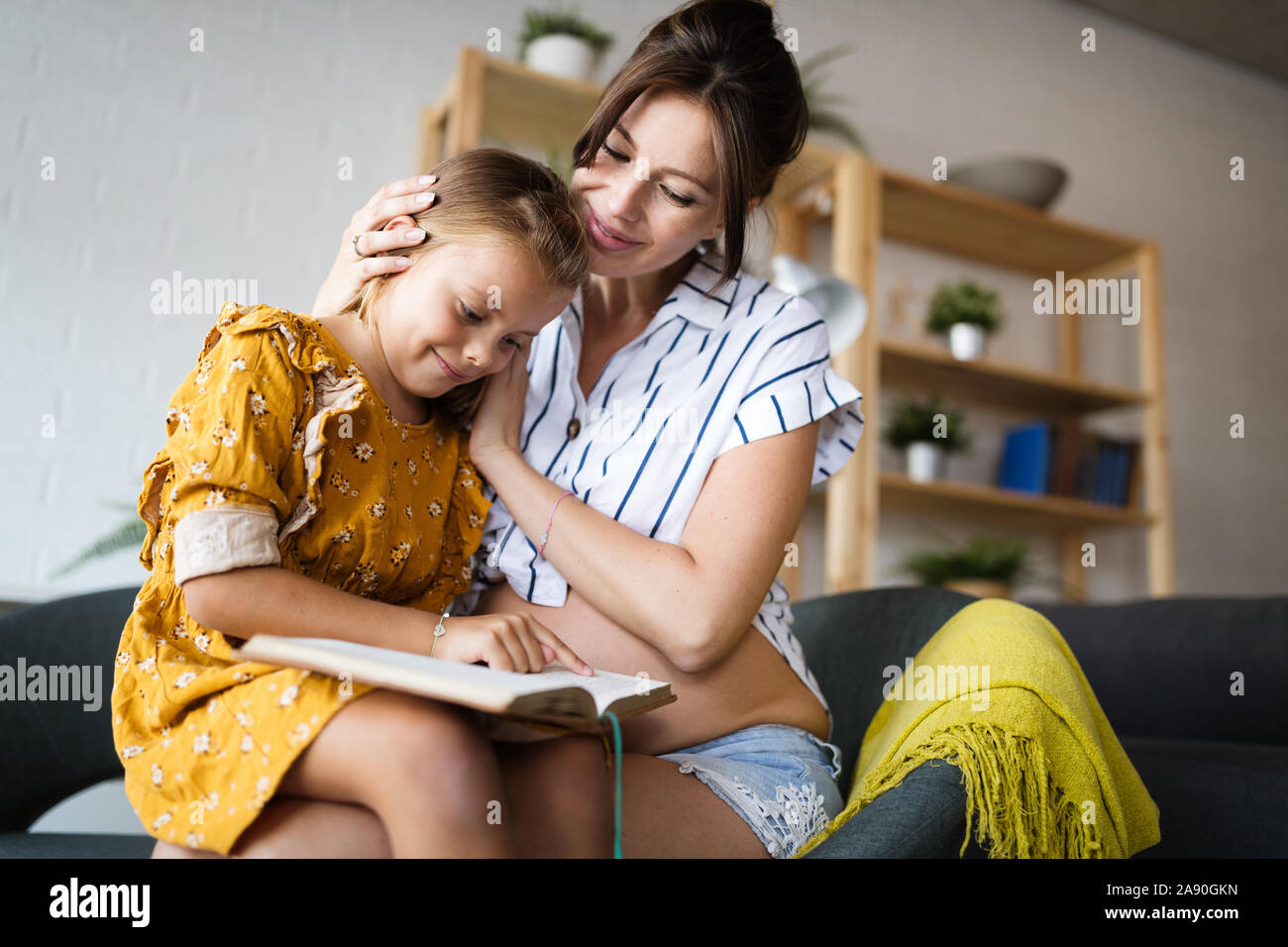 Portrait of pregnant mother and daughter reading a book in living-room Stock Photo