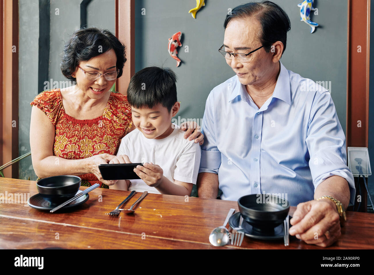 Happy Asian grandparents and grandchild sitting at cafe table and using