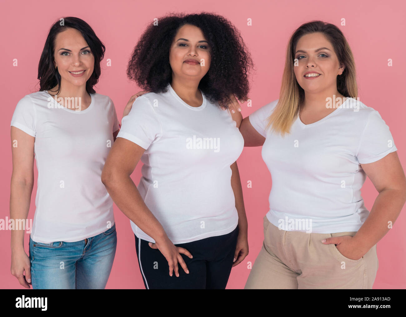 Three girlfriends smile beautifully and pose for the camera on a pink background. Girls in white t-shirts. Layouts for design Stock Photo