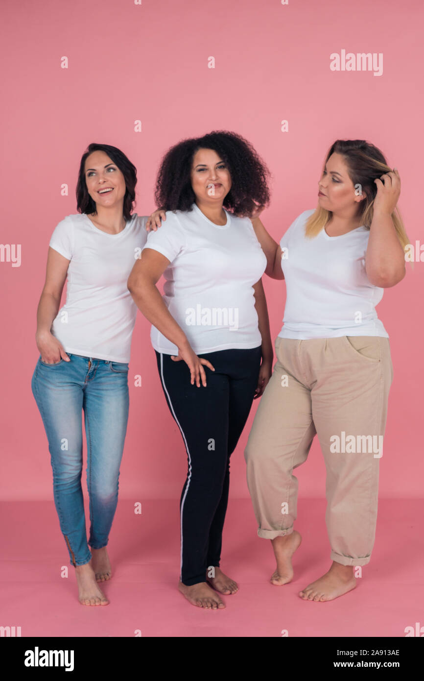 Three girlfriends smile beautifully and pose for the camera on a pink background. Girls in white t-shirts. Layouts for design Stock Photo