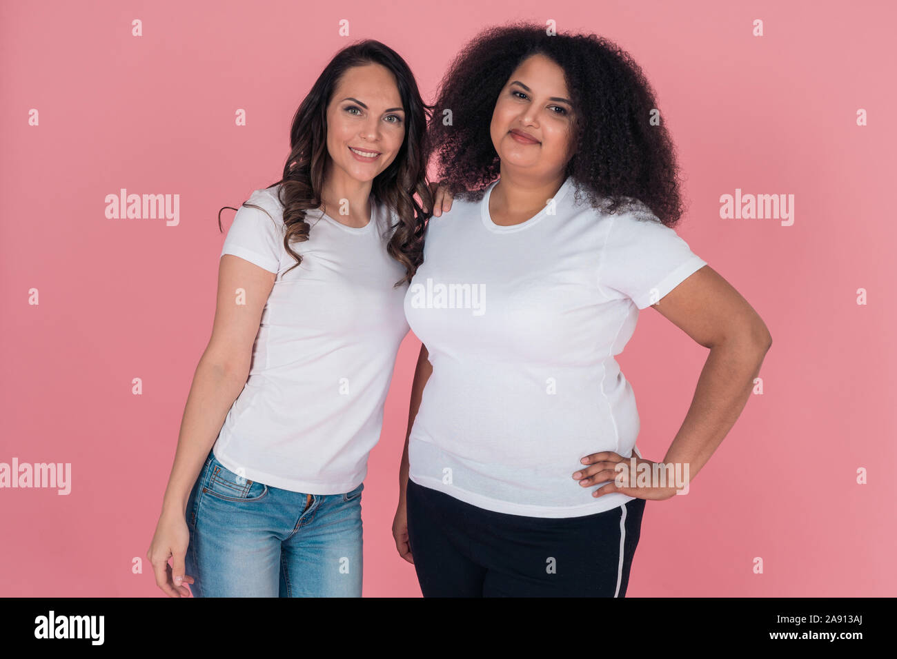 Two cute girls an African American and a Caucasian girl smile directly at the camera in white t shirts on a pink background Stock Photo