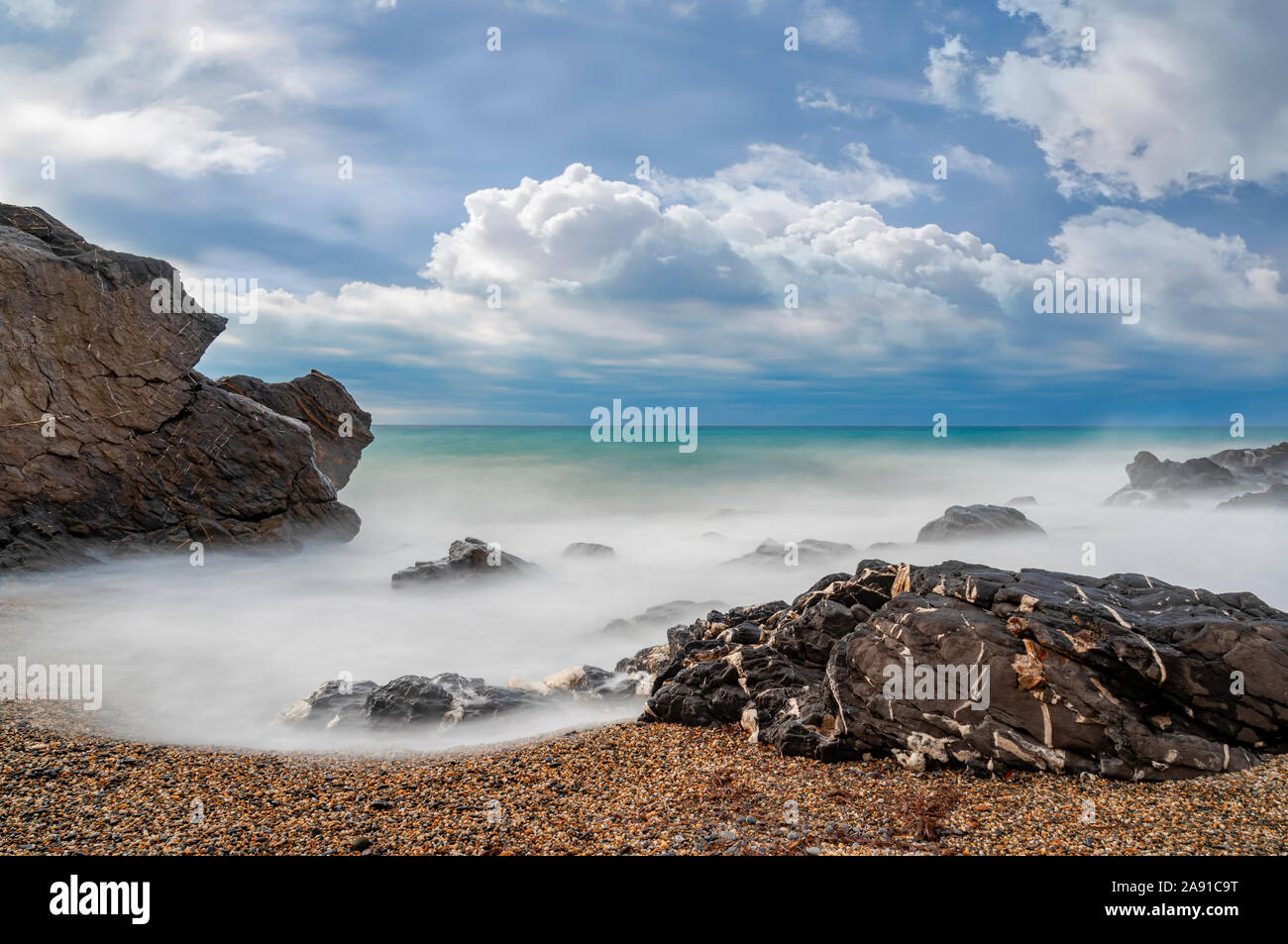 Long exposure of waves breaking on rocks on a sandy beach on the Mediterranean coast in Italy with white cumulus clouds in the sky Stock Photo