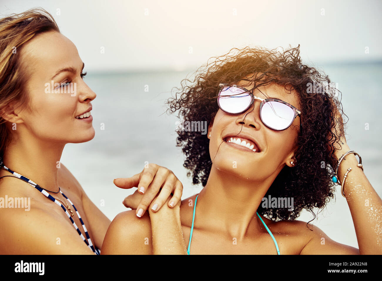 Two smiling young women standing together on a tropical beach while on summer vacation together Stock Photo