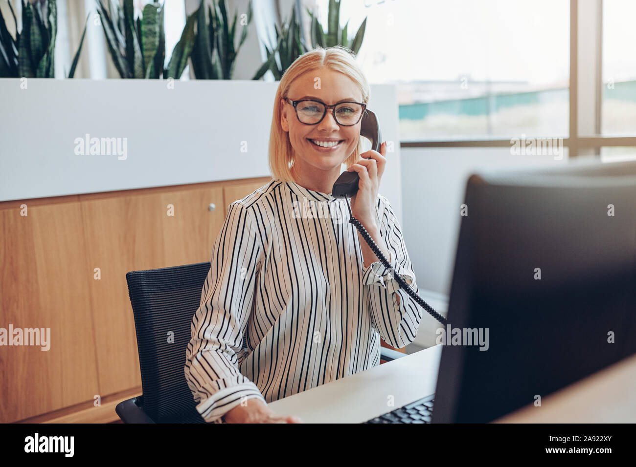 Smiling young businesswoman sitting at her desk in a bright modern office talking with a client on the telephone and working on a computer Stock Photo