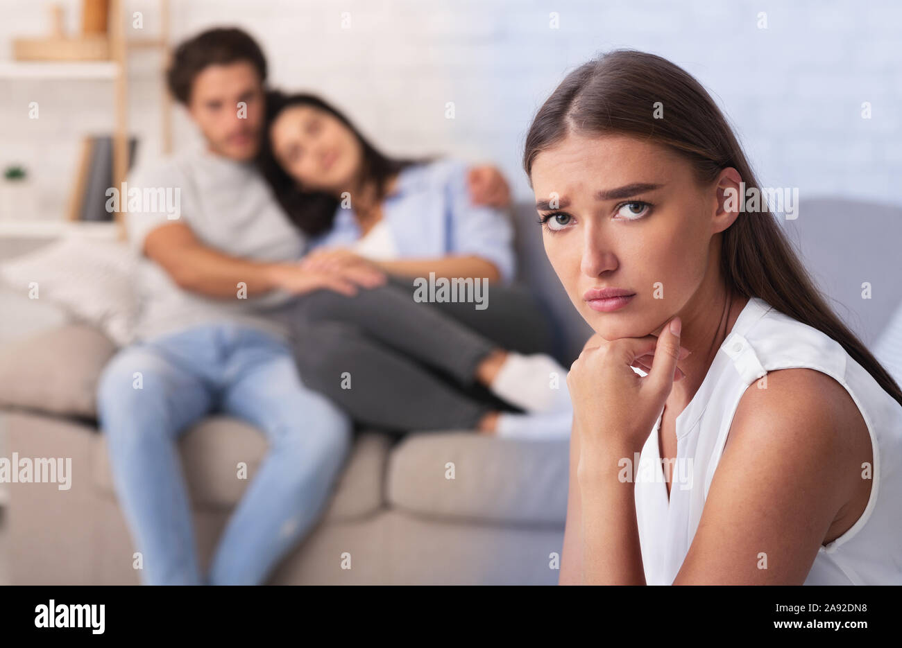Sad Woman Watching Couple Embracing Sitting On Couch Indoor Stock Photo