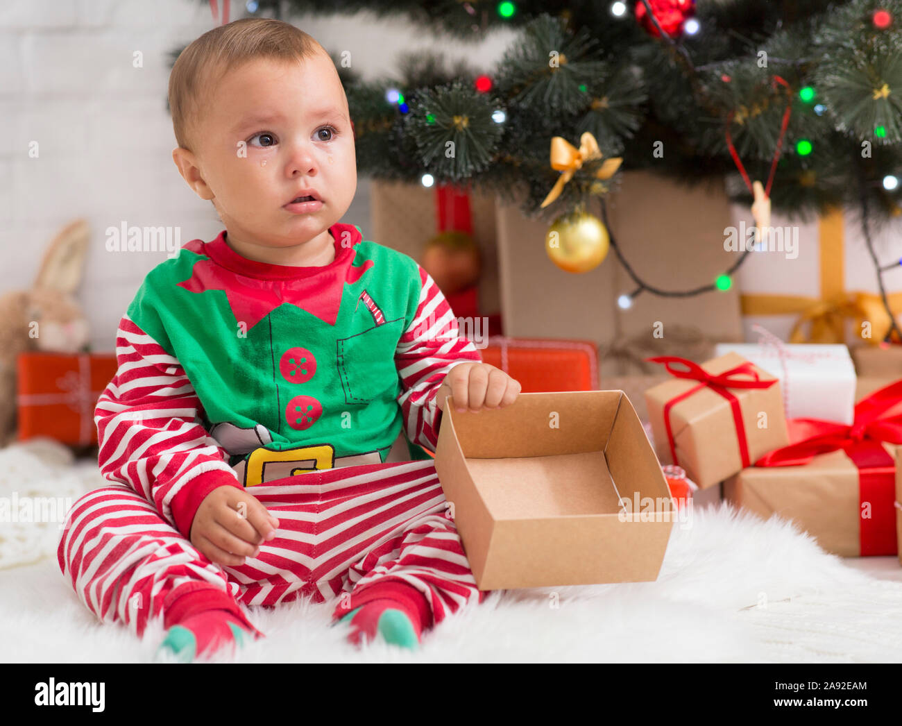 Unhappy baby elf with empty present box sitting near Xmas tree Stock Photo