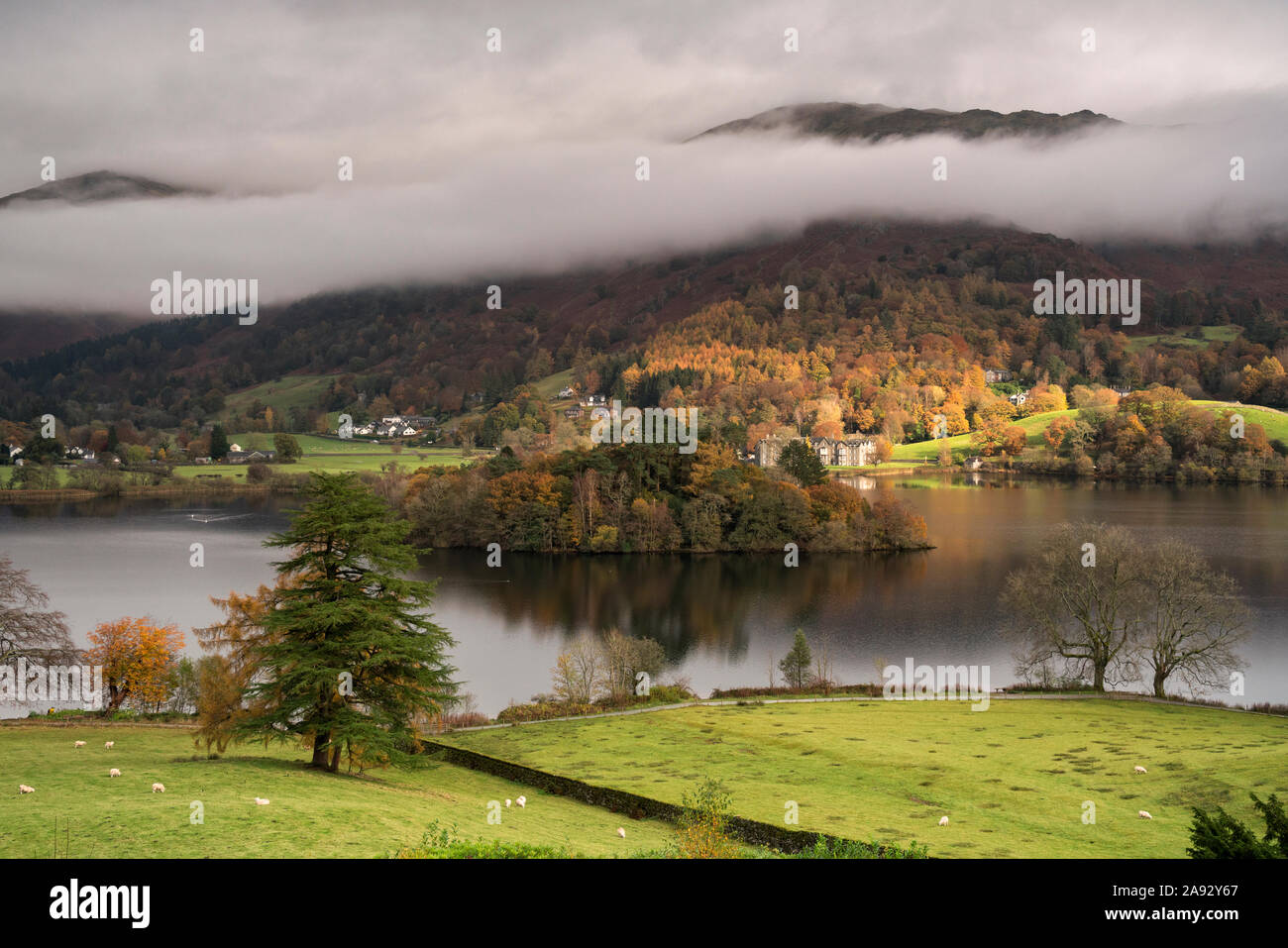 Sunshine breaking through low cloud and mist over Grasmere island in the middle of Grasmere lake on a rainy autumnal day Stock Photo