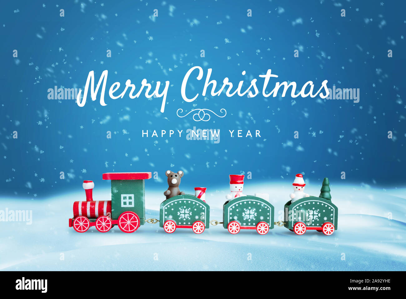 Cute wooden train in snow surrounded by a lot of snowflakes. Merry Christmas text on blue background. Christmas, New Year greeting card. Stock Photo