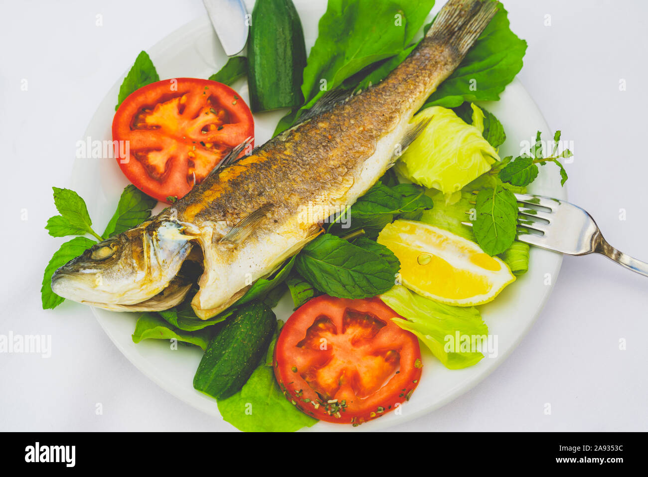 Grilled trout and spinach, mint, greens, lemon, tomato and cucumber on white plate with knife and fork on white background. Part of low-calorie diet Stock Photo