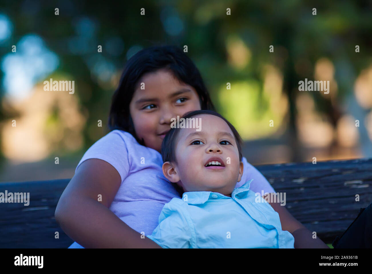 Younger brother leaning on her older sister as they watch a sunset on a park bench. Stock Photo