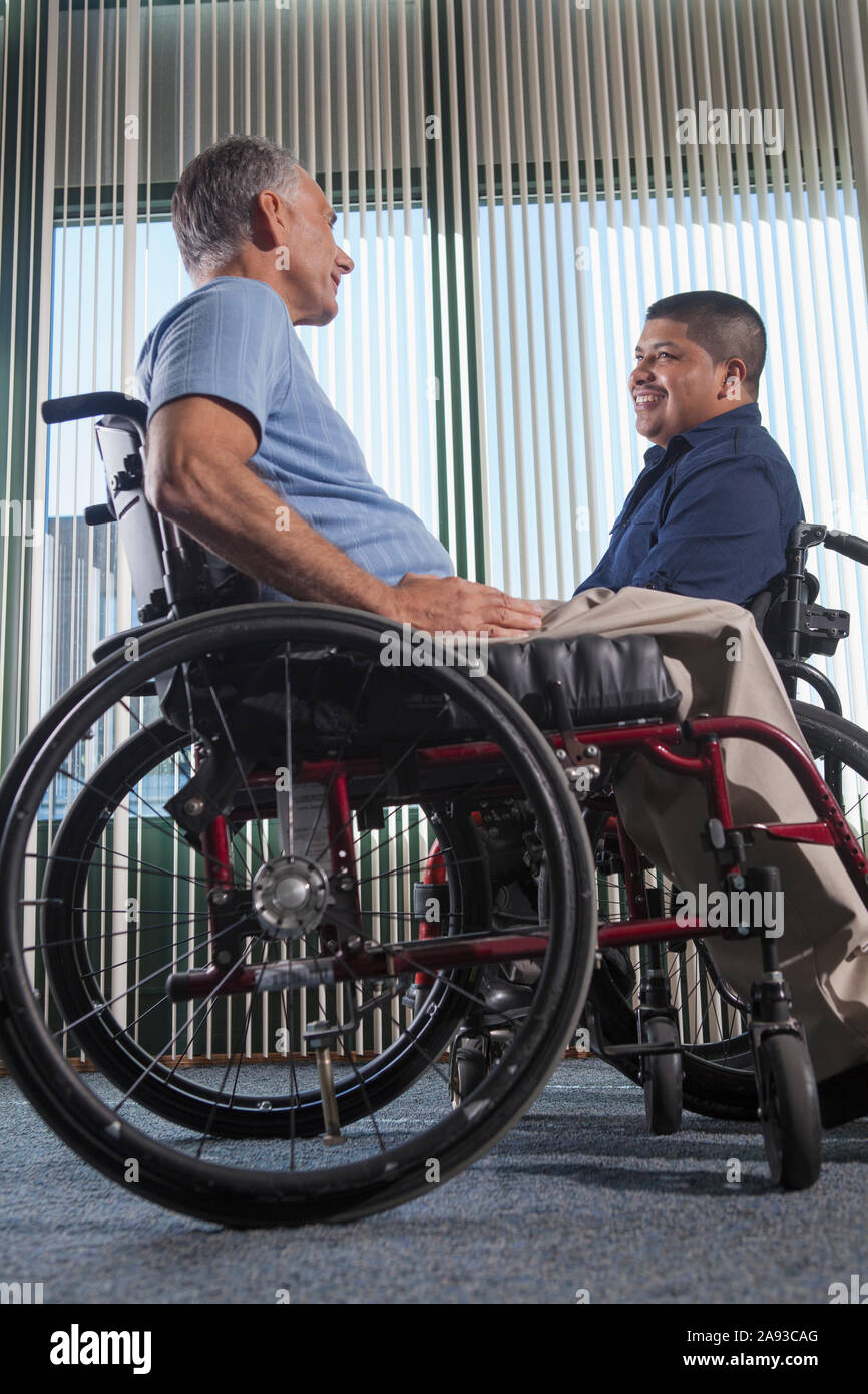 Two men with Spinal Cord Injuries on wheelchairs talking in an office corridor Stock Photo