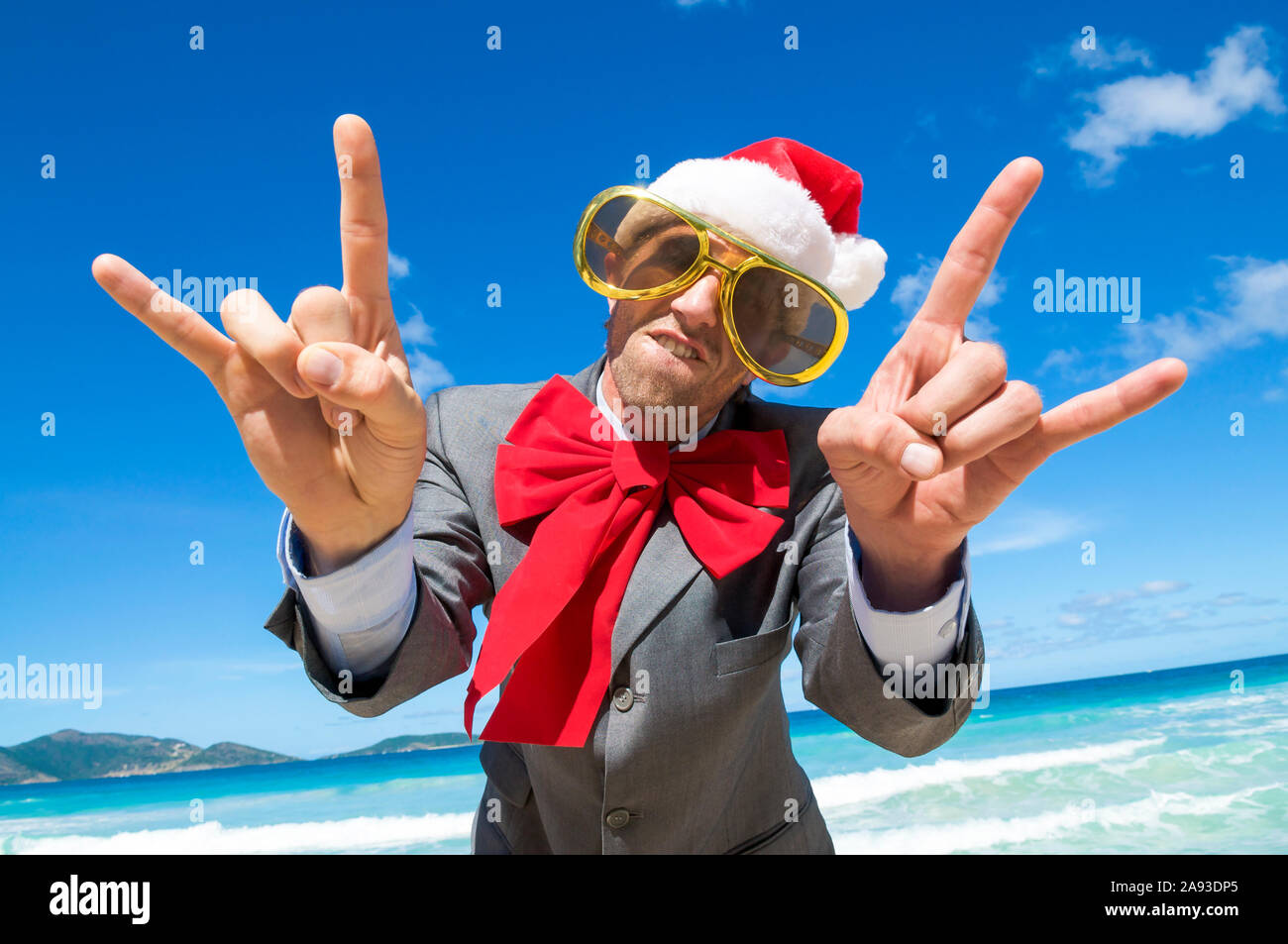 Funky businessman in Santa hat, sunglasses and big red Christmas bow making  rock n roll gestures on a tropical beach Stock Photo - Alamy