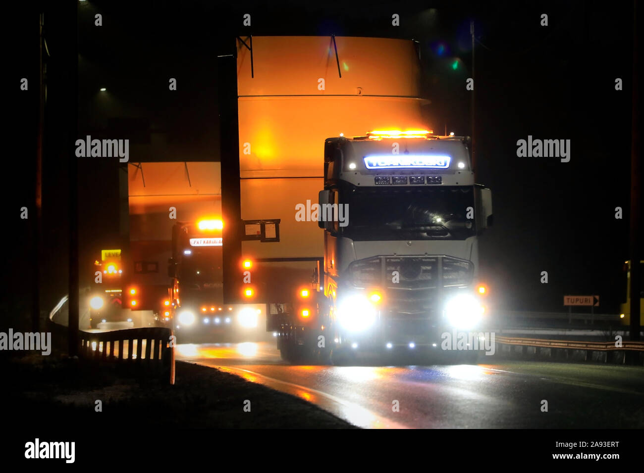 Convoy of two oversize load transports of a 5,5 m high steel frame with escort vehicles and warning lights at rainy night. Salo, Finland. Nov 8, 2019. Stock Photo