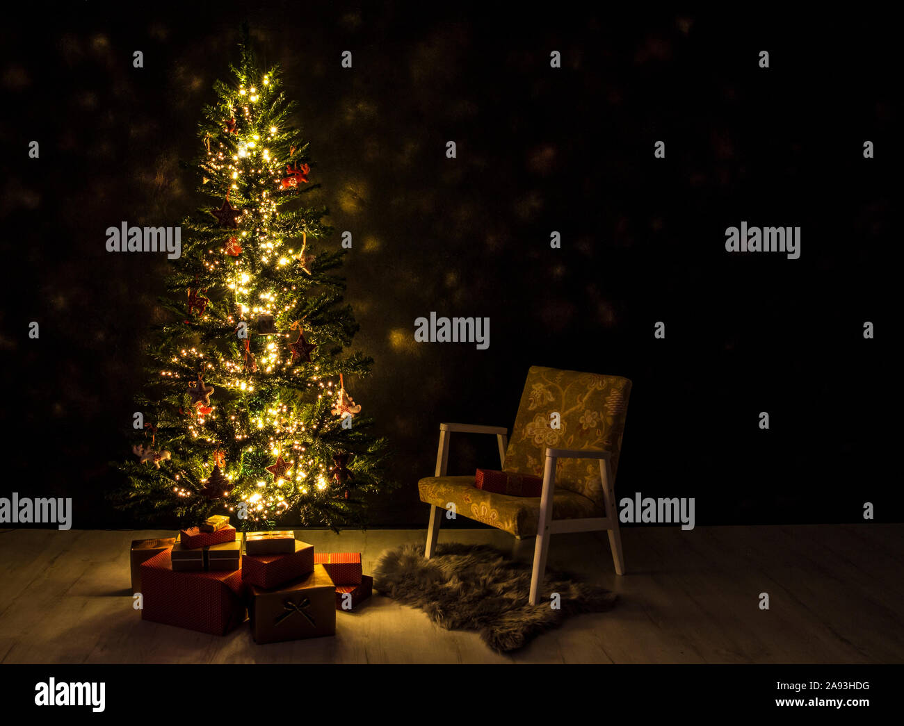 Decorated magical Christmas tree in front of black wall with Christmas red paper wrapped presents under the tree, retro chair next to it. Night. Stock Photo