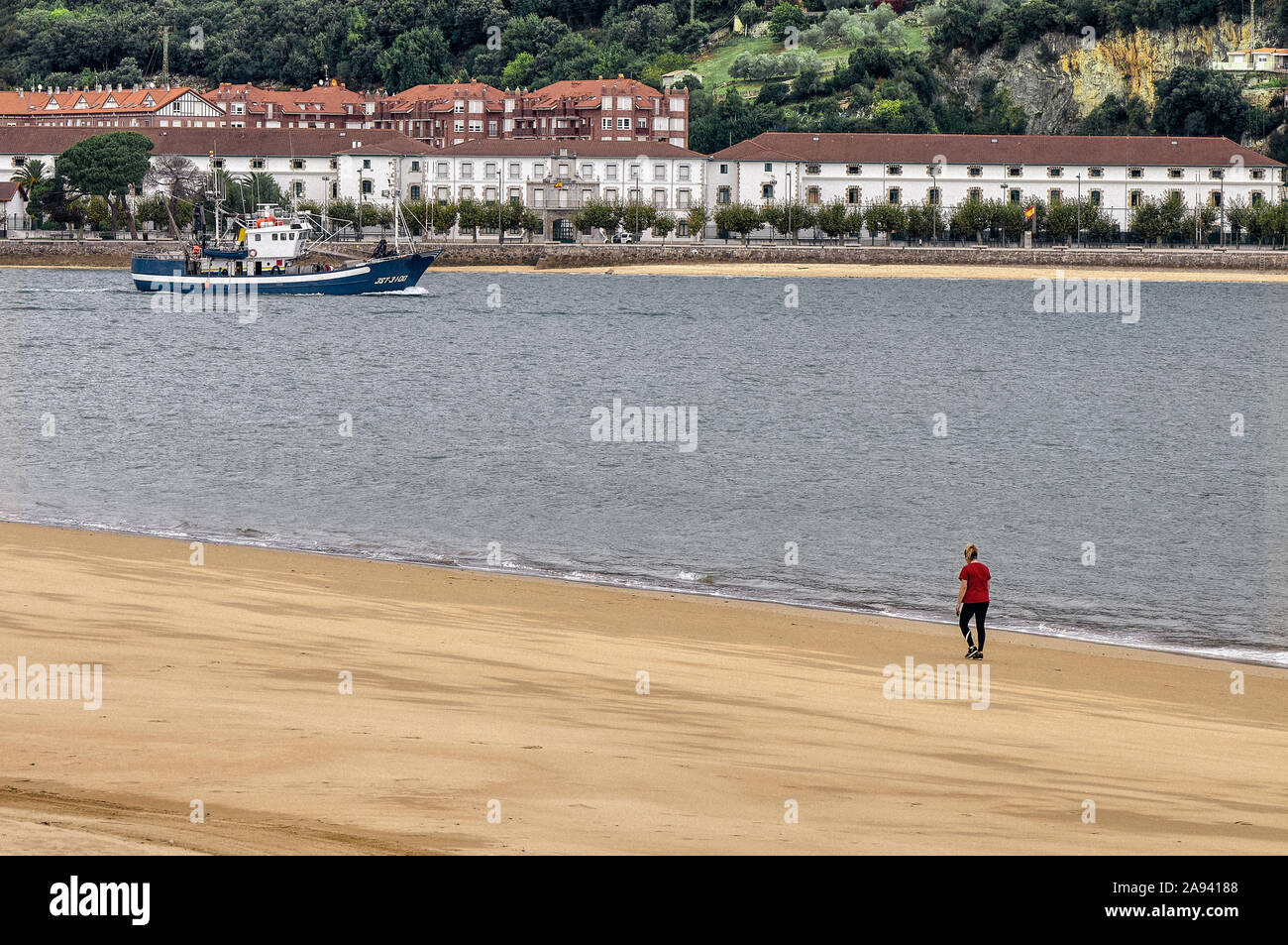 Woman walking along the seashore and a blue boat that goes fishing in the Bay of Biscay along the estuary of the Ason River, Laredo, Cantabria, Spain. Stock Photo