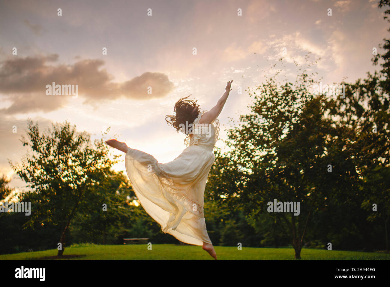Dancer leaps against cloud-filled sky in park at sunset in gold light Stock Photo