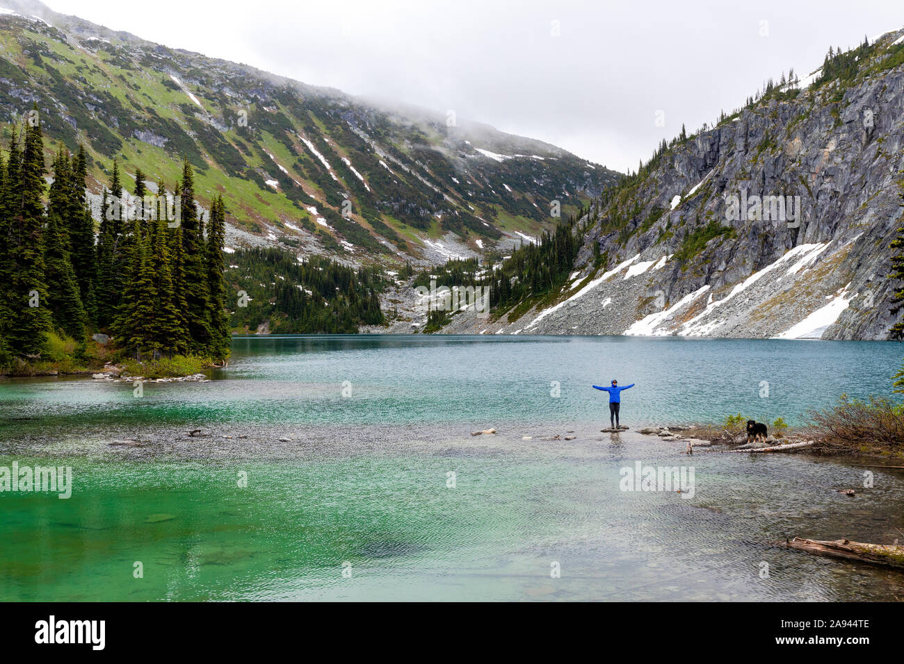A women hangs out on the side of an alpine lake with her dog and takes in the view in the Coast Mountains around Pemberton, British Columbia, on a rainy summer day. Stock Photo