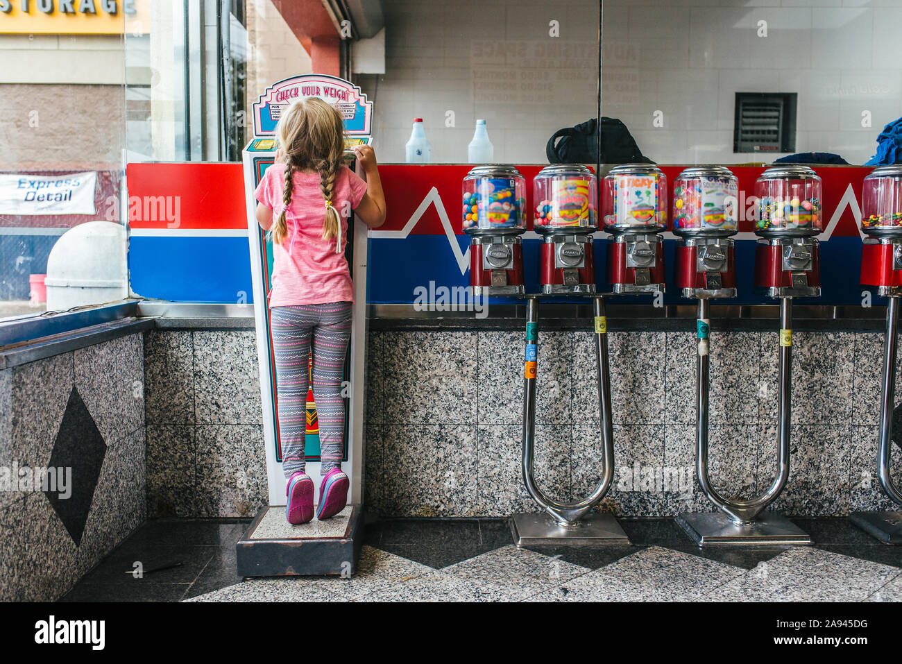 A girl stands on a scale at a car wash next to gumball machines. Stock Photo