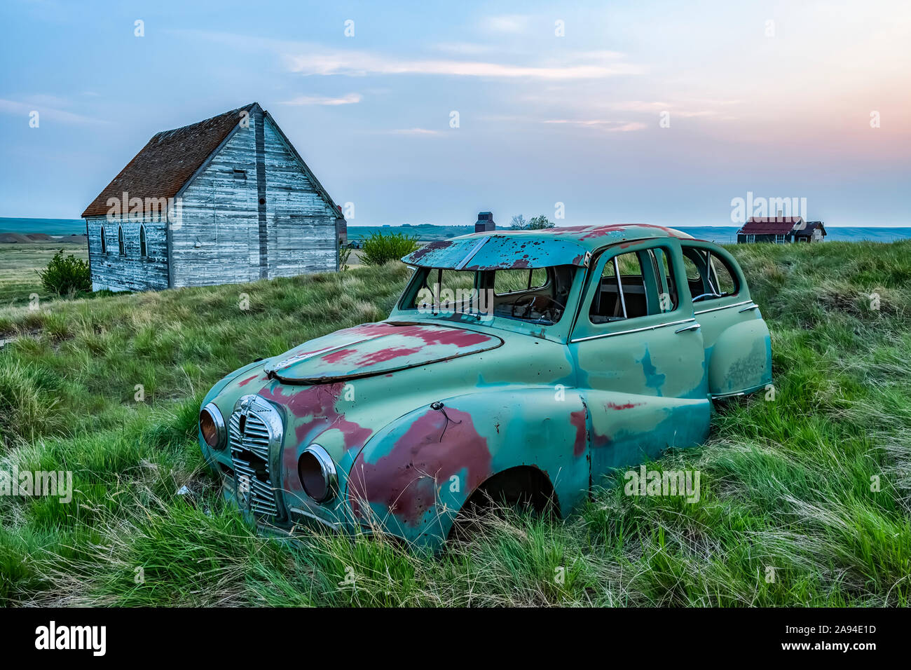 Vintage car sitting in the overgrown grass in a field with old buildings on a farmstead; Saskatchewan, Canada Stock Photo