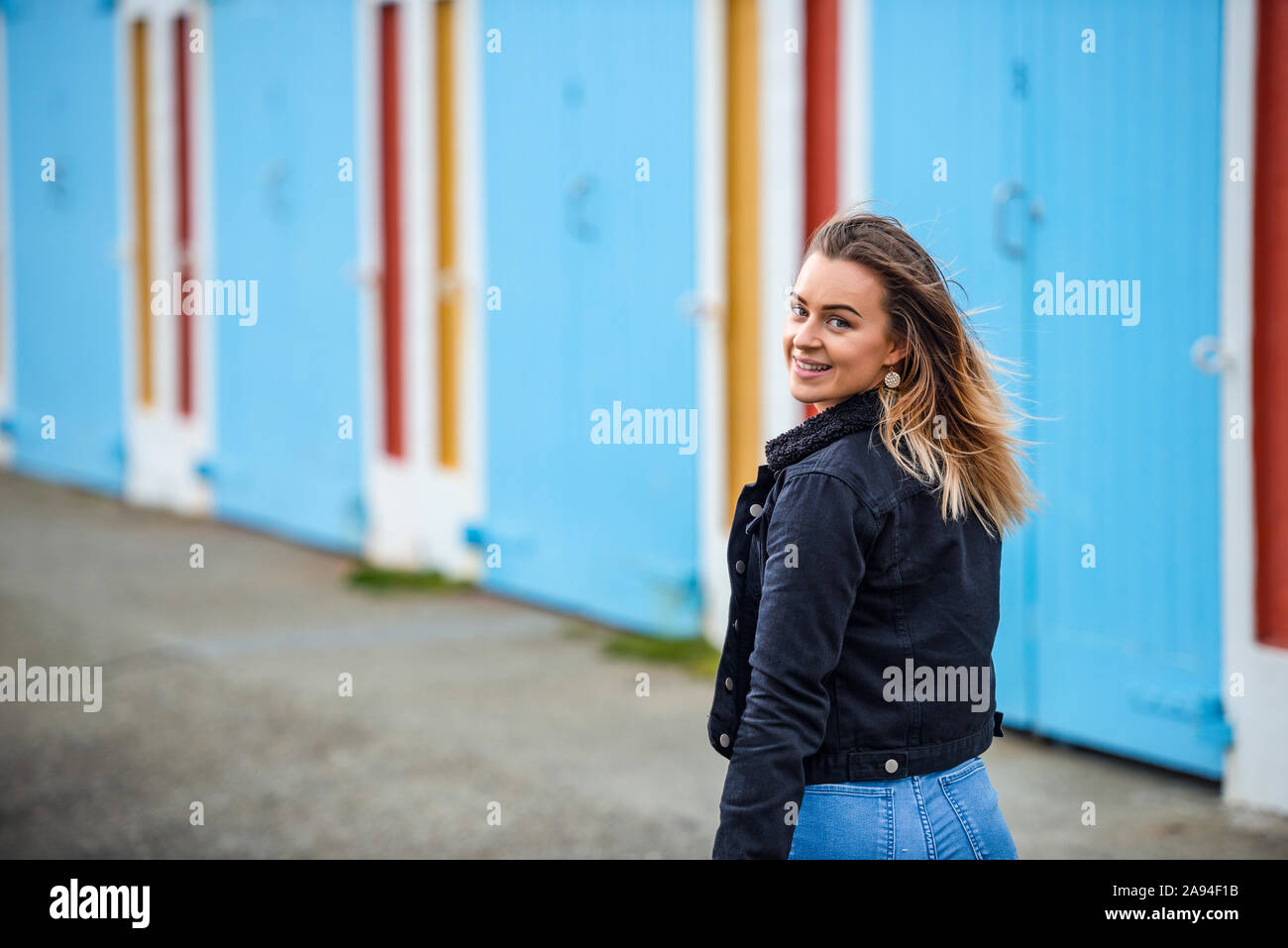 A young woman walks down the street along a colourful building, looking back over her shoulder at the camera; Wellington, North Island, New Zealand Stock Photo