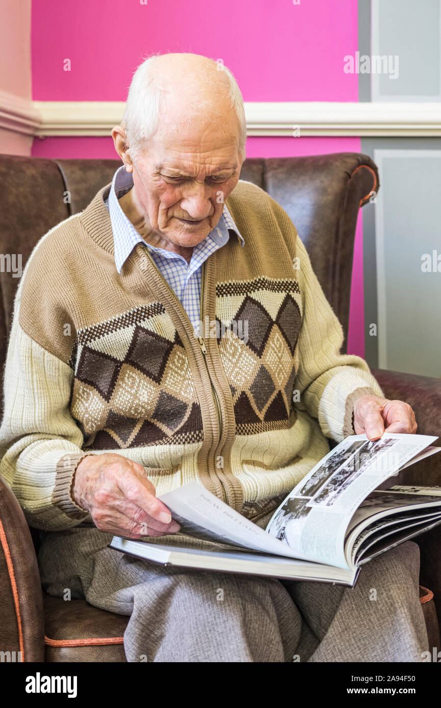Senior man sitting in a chair looking at a book about World War 2; Hartlepool, County Durham, England Stock Photo