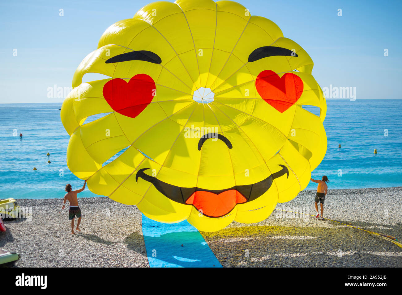 Two young men guide a happy face parasail parachute as it leaves the beach at the Bay of Angels on the Riviera in Nice, France. Stock Photo